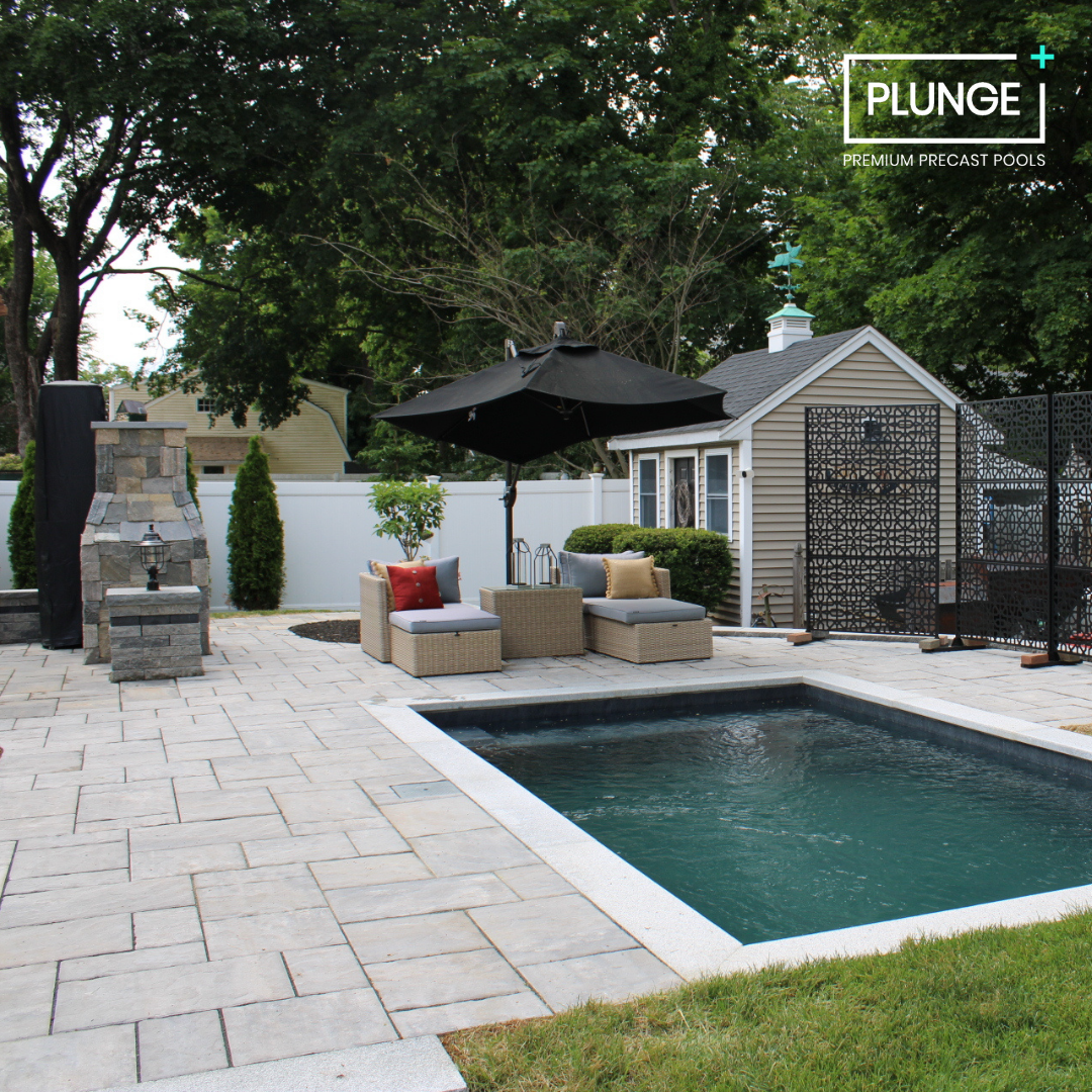 Backyard with a small pool, outdoor seating with cushions and a large black umbrella, stone patio, white fence, small shed, and trees in the background.