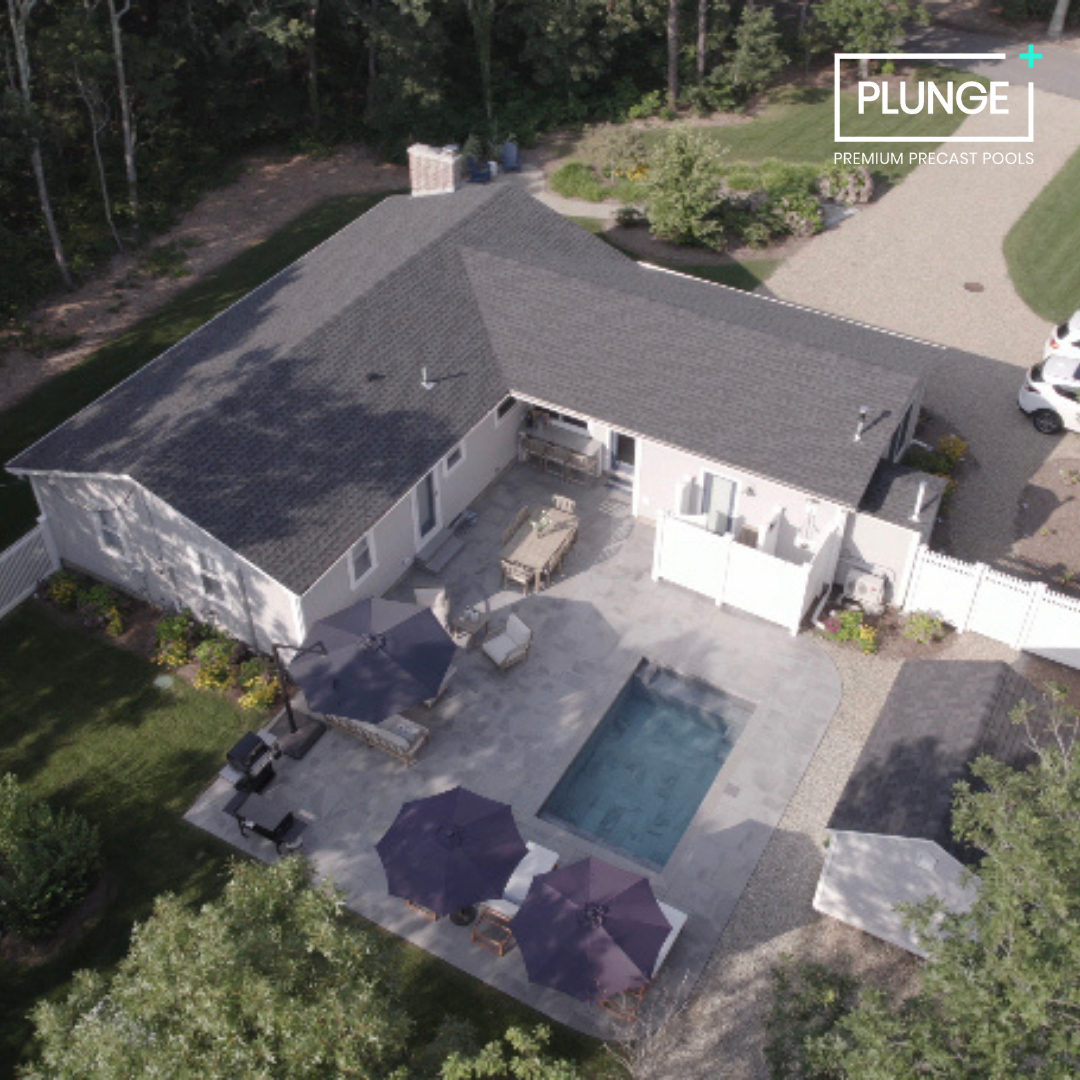 Aerial view of a backyard with a swimming pool, patio furniture, and umbrellas, attached to a house with a gray roof. There are trees and a driveway with parked cars nearby.
