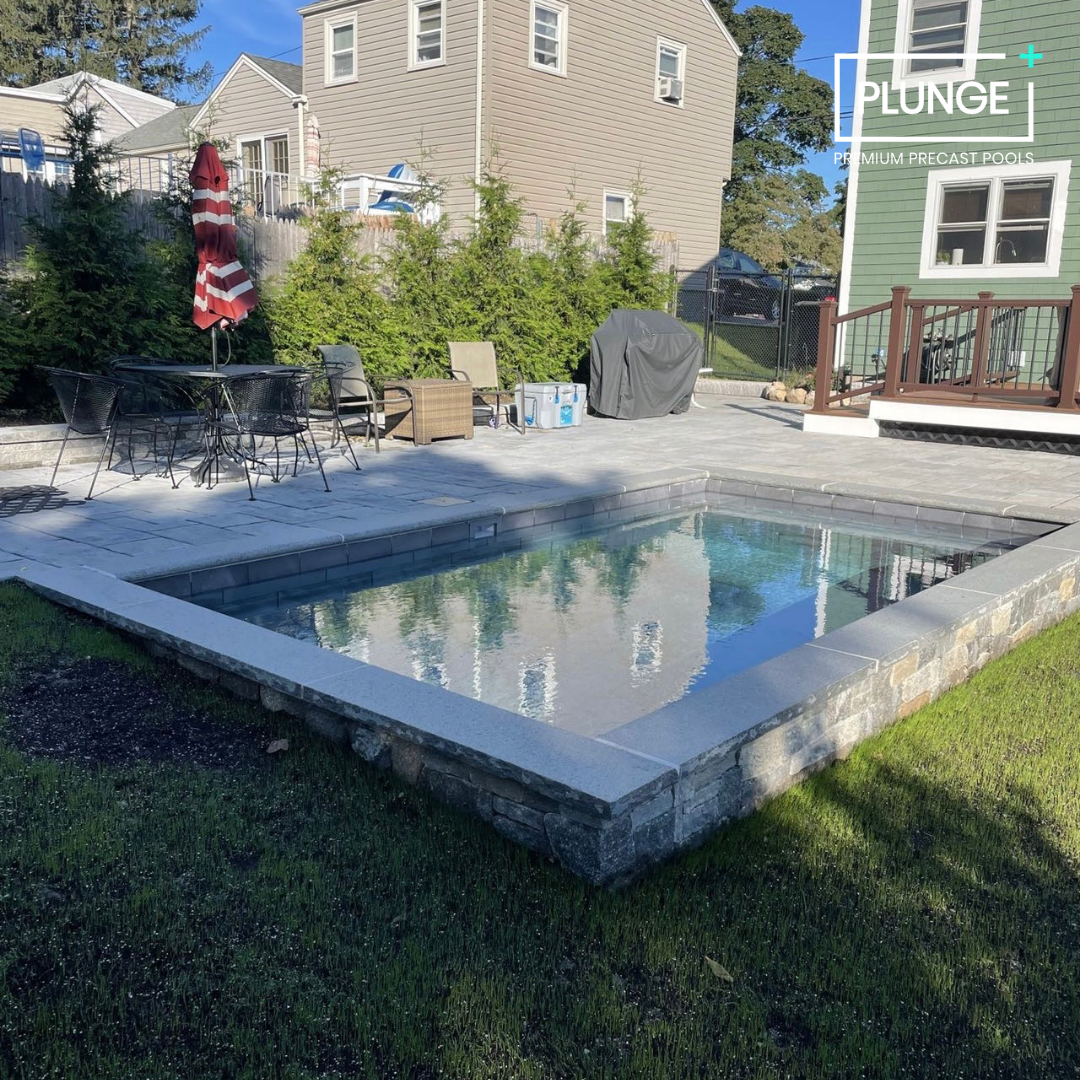 Residential backyard with a small pool, outdoor seating including a table with a red and white umbrella, a barbecue grill covered with a black cover, and two chairs. There are some coolers and chairs on the patio, with a house and trees in the backgr