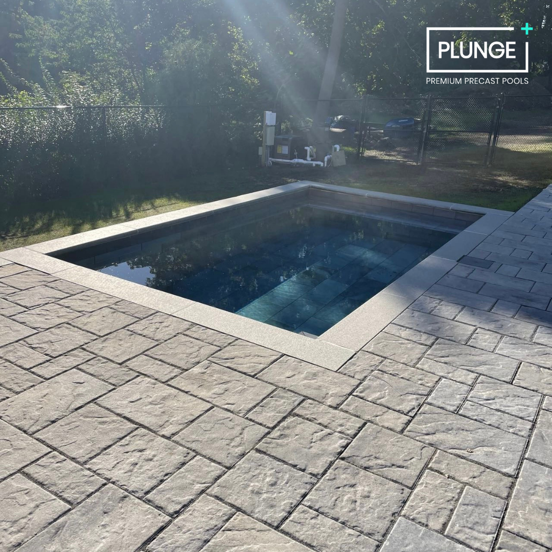 A rectangular inground pool with a stone border and concrete paving surrounding it, located outdoors with trees and a fence in the background.
