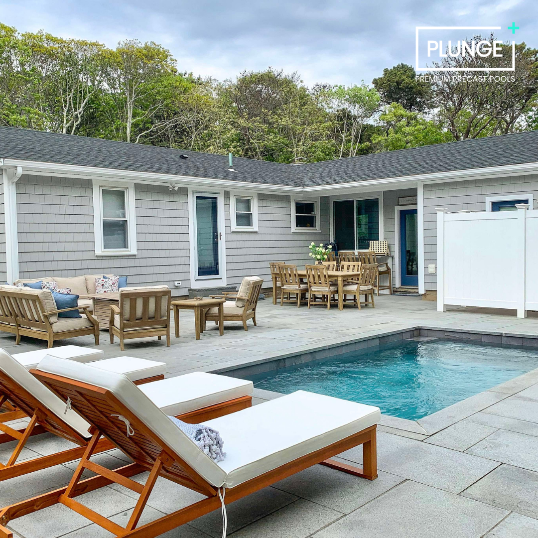 Outdoor backyard with a small swimming pool, wooden lounge chairs with white cushions, a seating area with wooden chairs and table, surrounded by a gray house with windows and doors, and a backdrop of green trees under a cloudy sky.