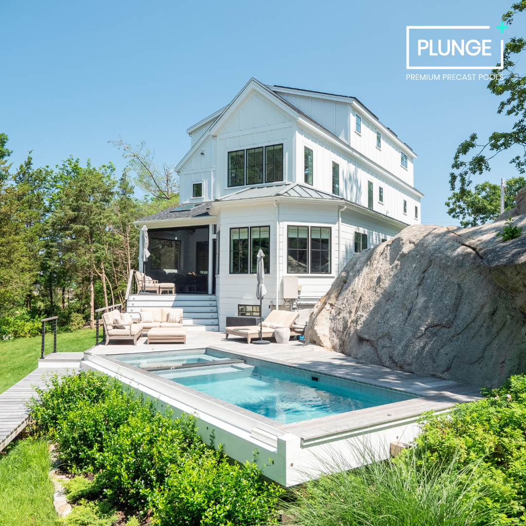 A modern white house with a backyard pool area, surrounded by greenery and a large rock, under a clear blue sky.