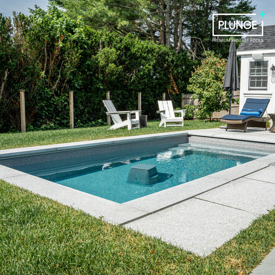 A backyard with a small in-ground pool surrounded by concrete paving and green grass. There are two white Adirondack chairs and a wicker lounge chair with a blue cushion and striped pillow. A black patio umbrella and a small shed or garage are also v
