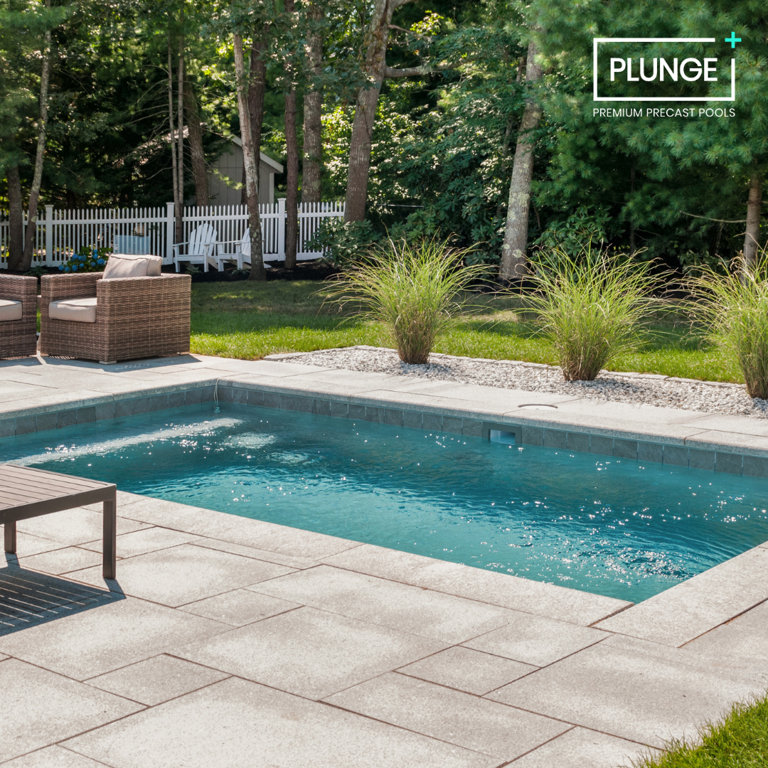Backyard with a small swimming pool, patio furniture, green plants, trees, and a white picket fence in the background.
