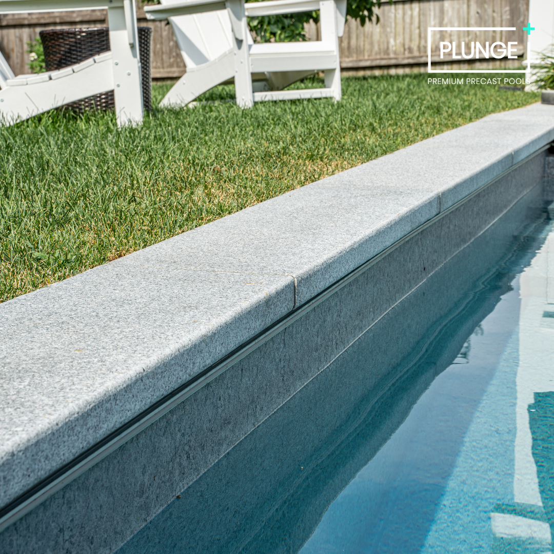 Close-up of a swimming pool with a grey stone edge, adjacent to a grassy yard with outdoor furniture and a wooden fence in the background.