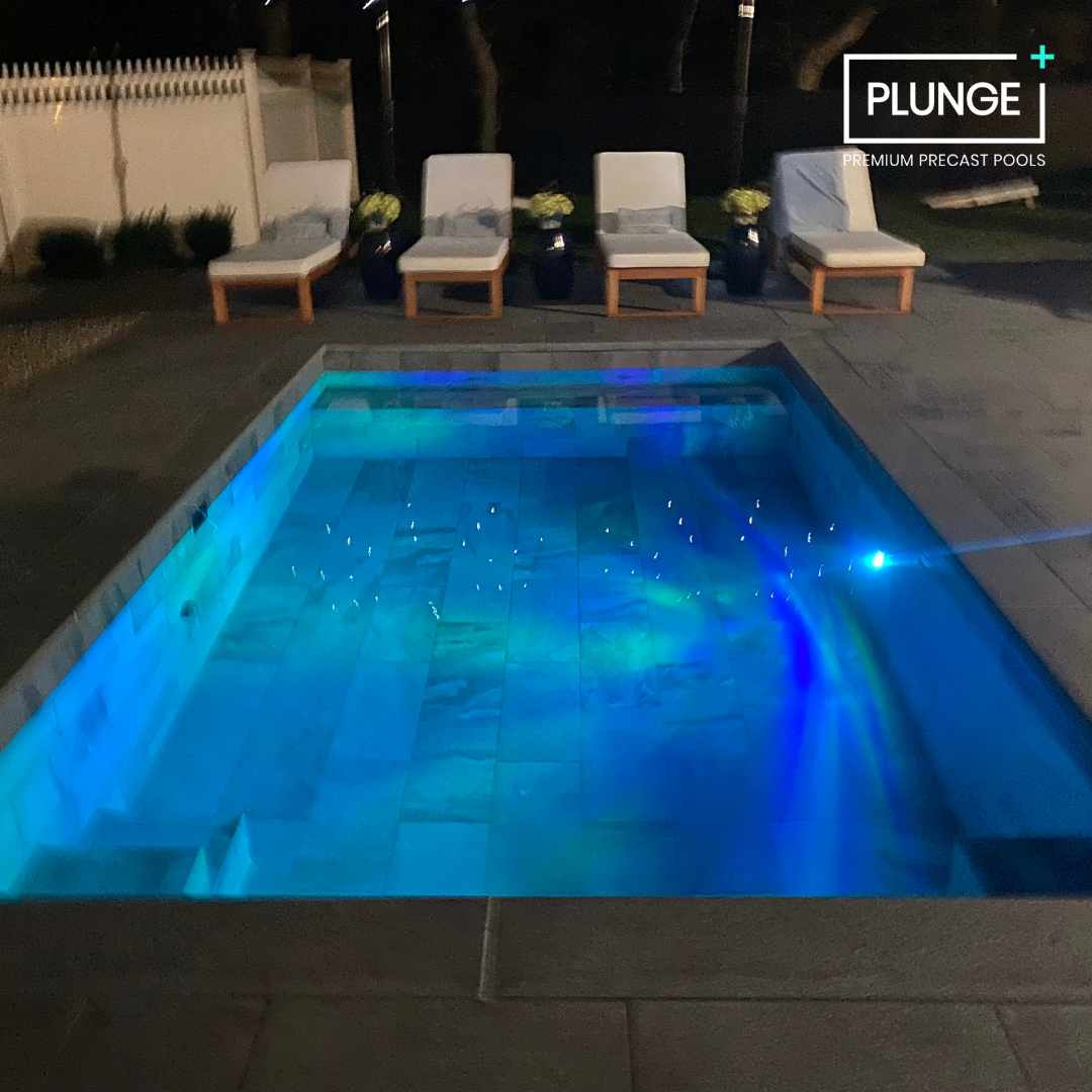 Nighttime view of a small illuminated swimming pool with blue LED lights, surrounded by four lounge chairs with cushions and potted plants, and a privacy fence in the background.