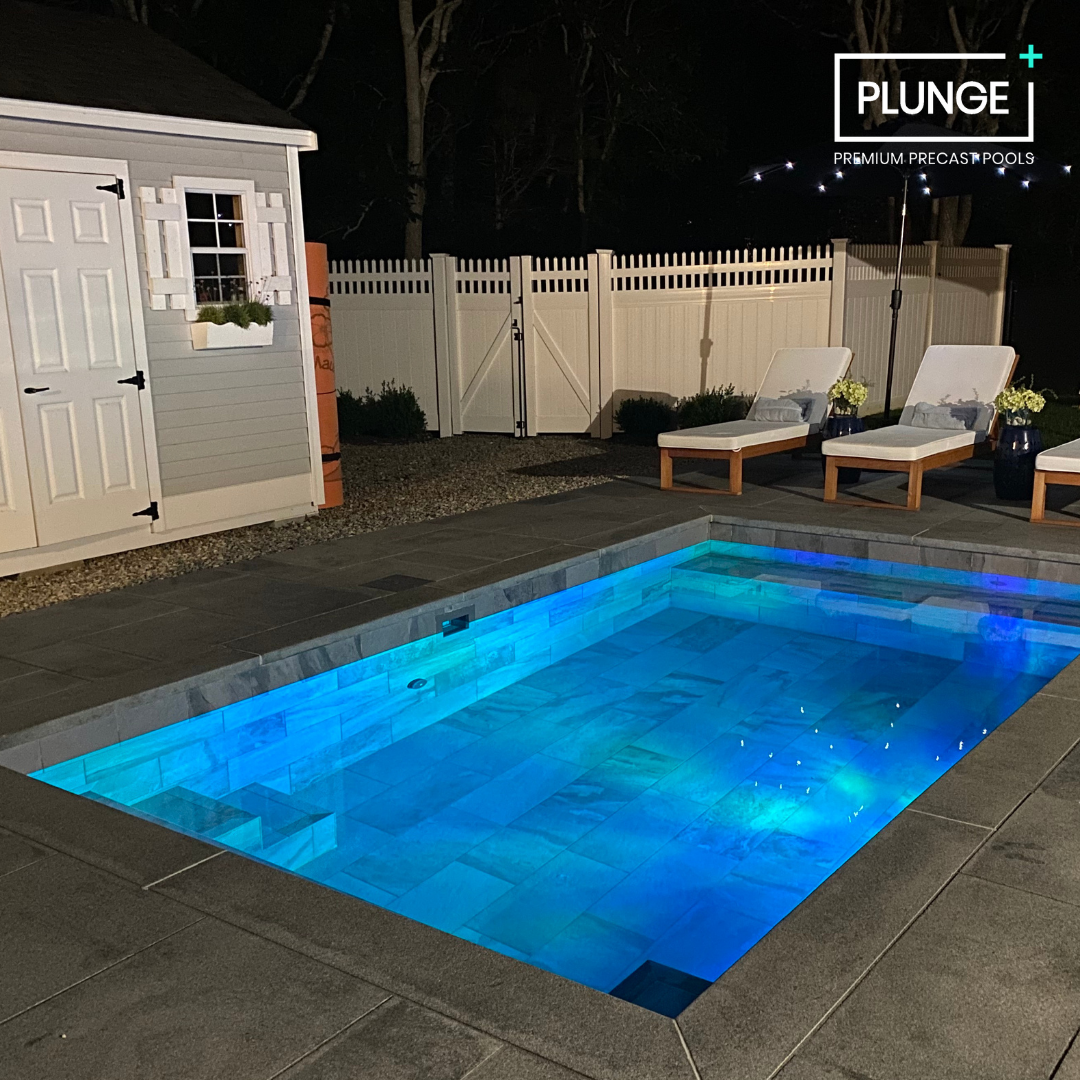 Night view of a backyard swimming pool illuminated with blue lights, surrounded by a concrete deck with lounge chairs and a small shed, enclosed by a white picket fence.