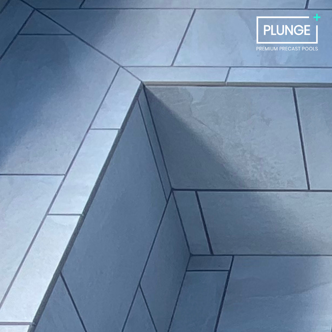 Close-up of a corner of an indoor pool with gray tiles and a step, with a logo in the upper right corner that reads 'PLUNGE Premium Precast Pools.'