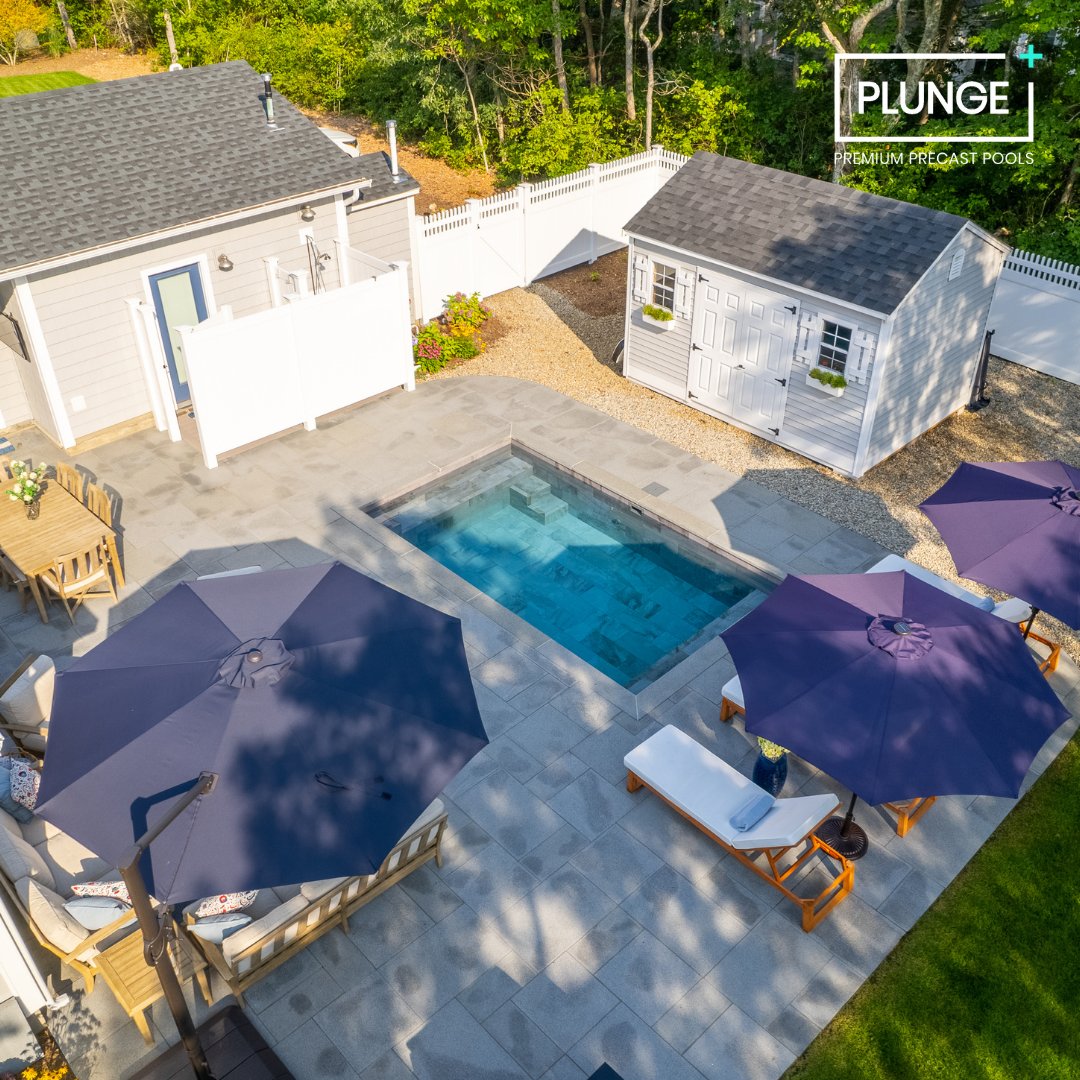 A backyard with a small rectangular swimming pool, outdoor furniture with purple umbrellas, a white shed, a house, and a white fence, surrounded by trees.