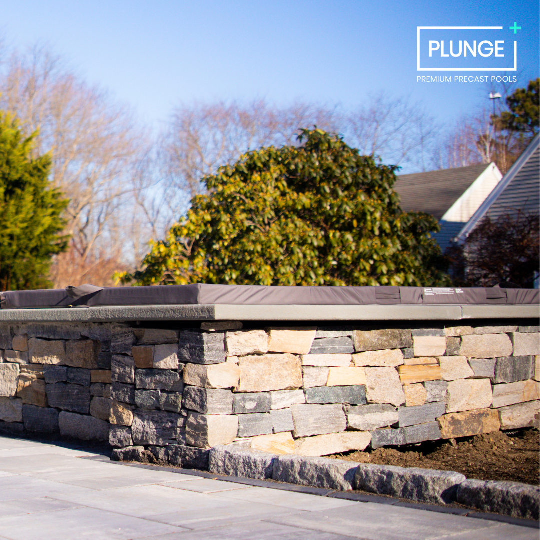 A backyard with a stone raised platform or wall, topped with a gray concrete cap, adjacent to a paved patio. In the background, there are leafless trees, a large green shrub, and a house with a sloped roof. The image features a logo for Plunge premiu