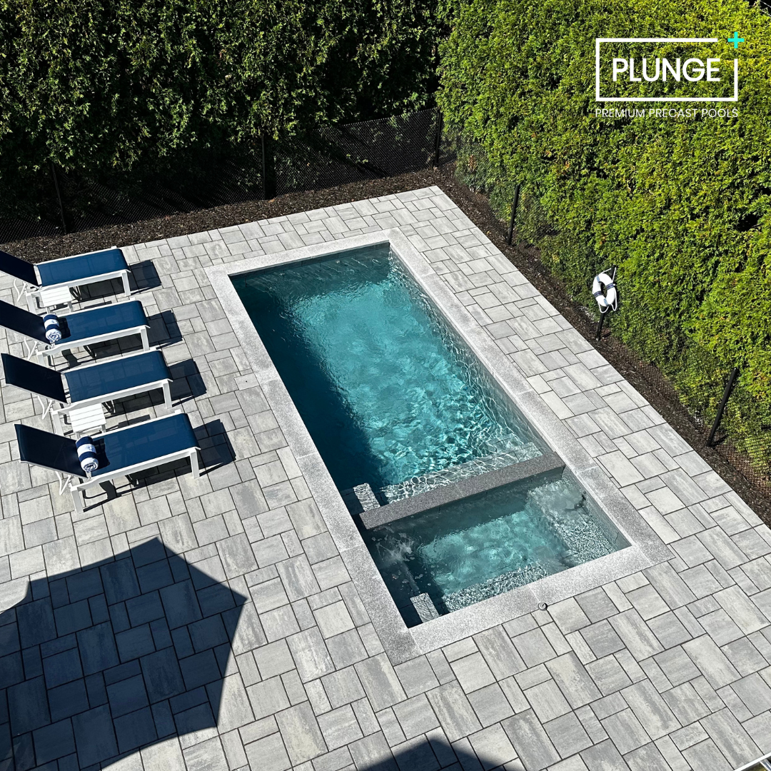 A rectangular outdoor swimming pool with a separate smaller hot tub, surrounded by a tiled deck, four blue lounge chairs with towels, a life ring on the hedge, and lush green hedges in the background.