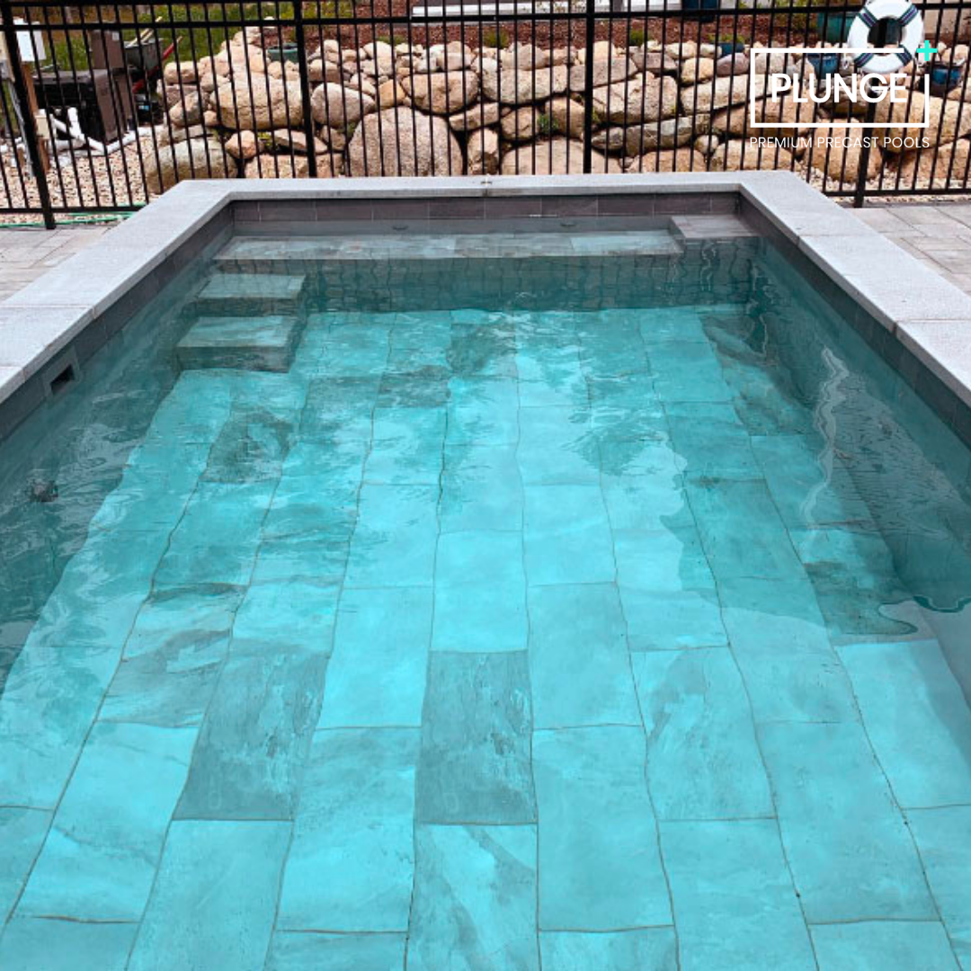 A rectangular private pool with clear water, light blue tiles, built-in steps at one end, and a stone border. A black metal fence surrounds the yard, with rocks and a rocky garden in the background.