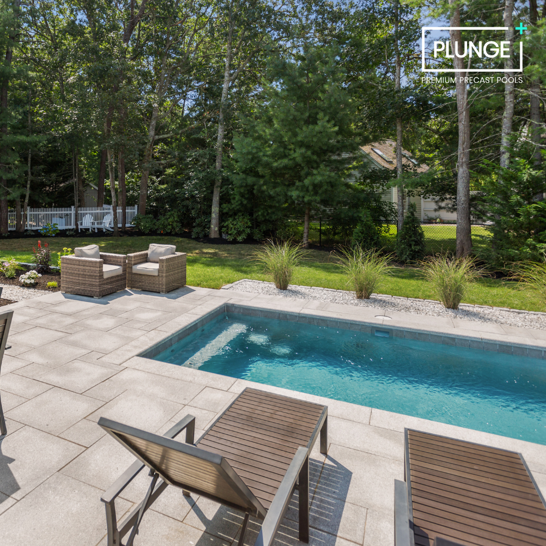 A backyard with a swimming pool, outdoor seating including two lounge chairs, and a small patio area with wicker chairs and a patio table. There are trees and a garden with flowers and ornamental grasses, and a house with a white fence in the backgro
