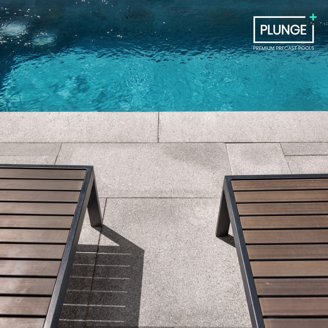A swimming pool with clear blue water, two lounge chairs on a stone-paved deck, and a logo in the top right corner that reads 'PLUNGE PREMIUM PRECAST POOLS'.