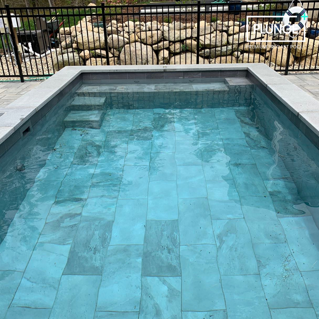 A small in-ground swimming pool with clear blue water, gray stone coping, and steps leading into the pool. Metal fence and rocks with some greenery are visible in the background.