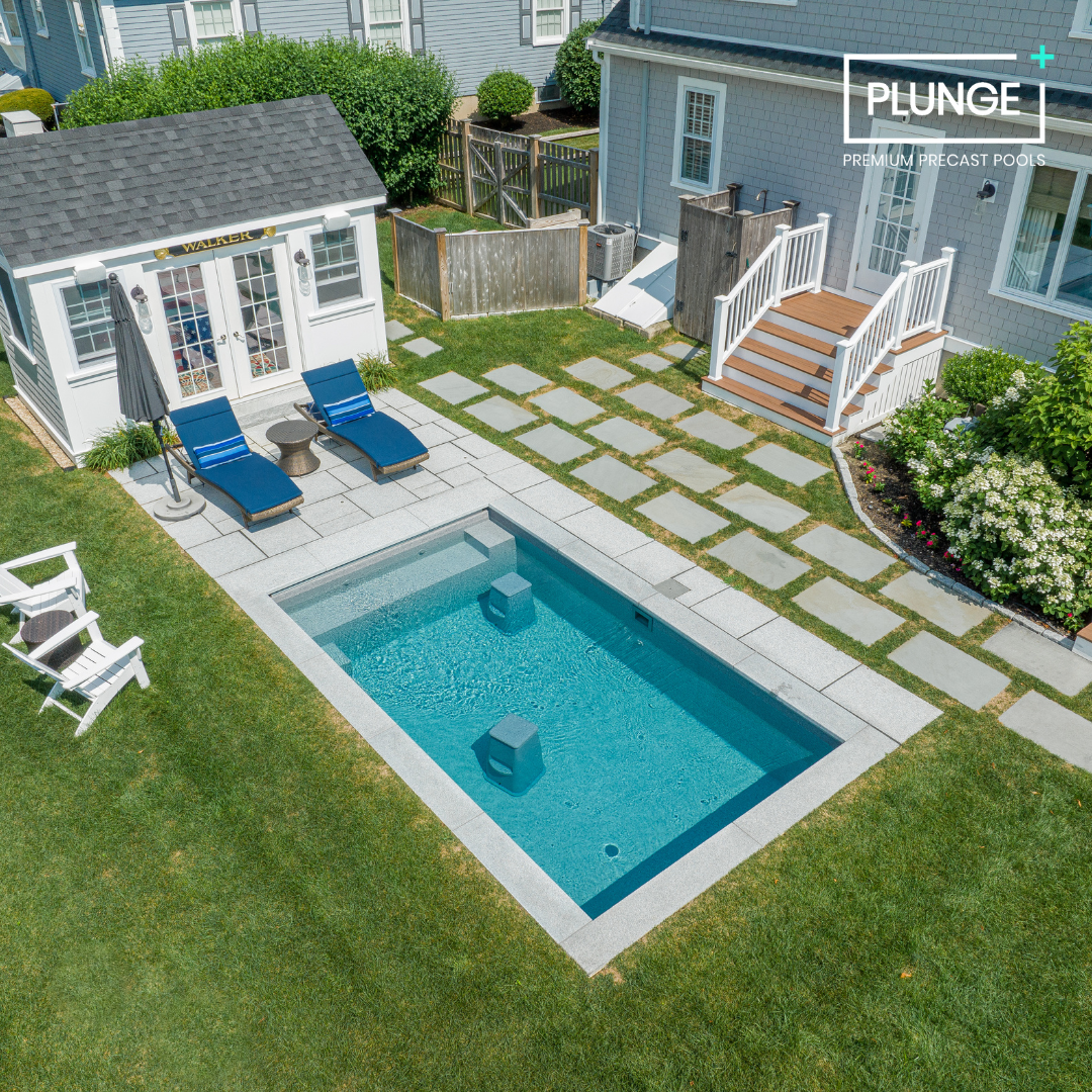 A backyard with a small rectangular swimming pool, two blue lounge chairs with a small table and umbrella, a white shed with a sign that says "Walker," a paved stone pathway, a small wooden deck with stairs, surrounding greenery, and a fence.