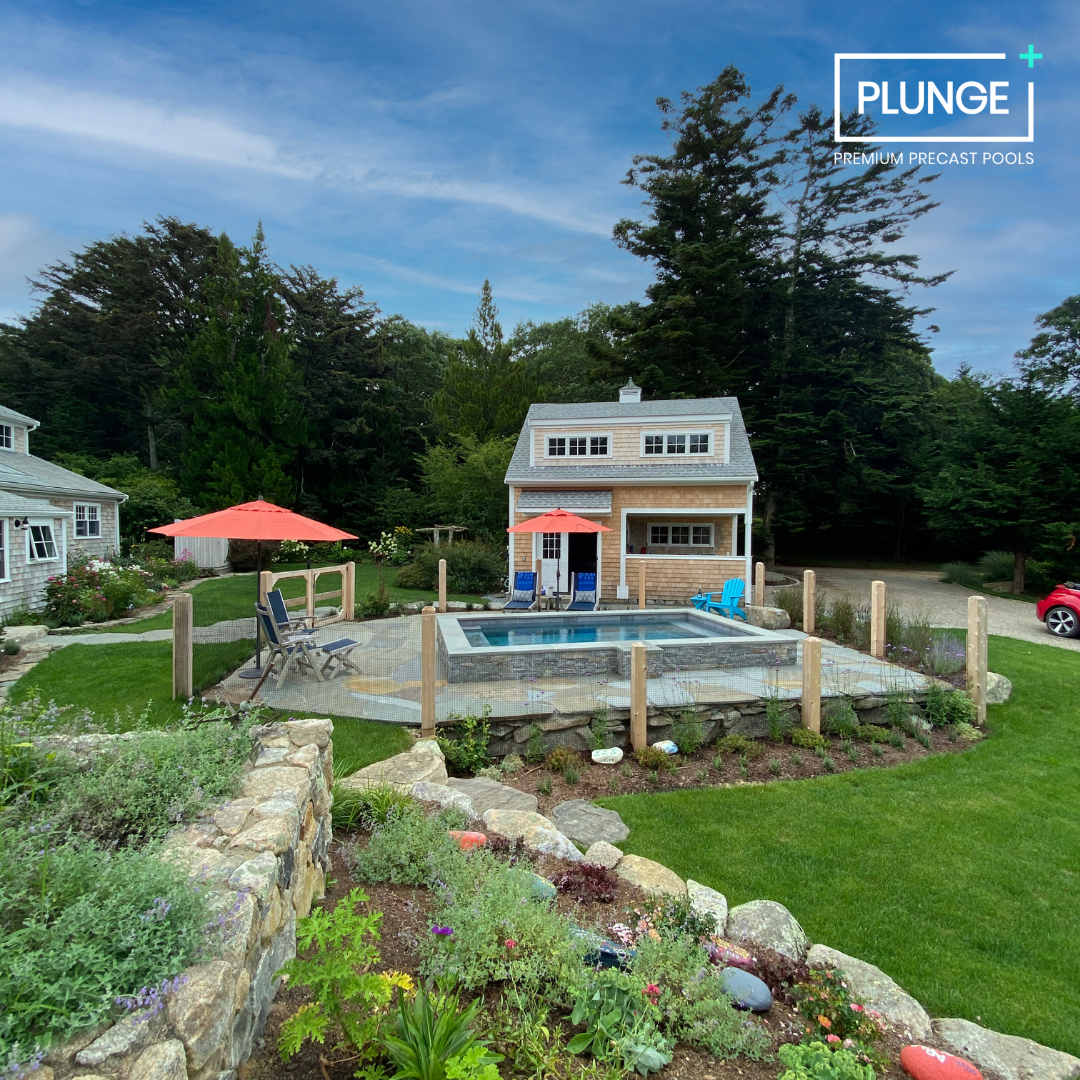 Backyard scene with a small pool, deck chairs, and umbrellas in front of a cottage-like house surrounded by trees and greenery.