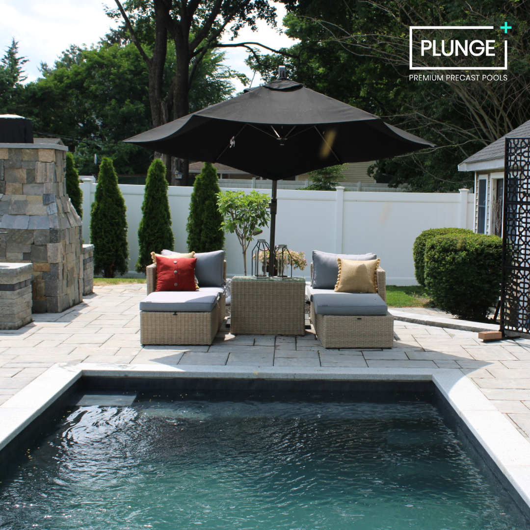 Outdoor backyard with a small swimming pool, patio furniture with cushions and pillows, a large umbrella, a stone fireplace, green shrubs, and a white fence in the background.