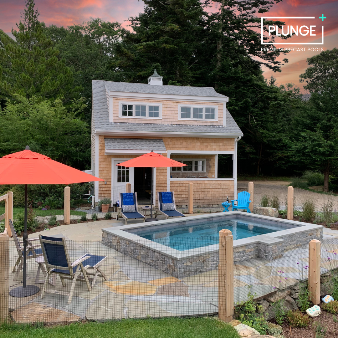 A backyard scene featuring a small house with a windowed upper level, a covered porch, and a landscaped yard with a small rectangular pool surrounded by stone and a paved patio. There are two orange umbrellas, a blue Adirondack chair, and several lou