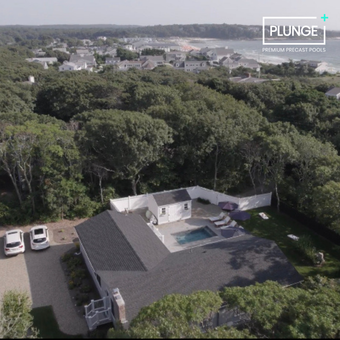A house with a gray roof and a backyard featuring a pool and patio umbrellas, surrounded by trees and neighboring houses in the distance.
