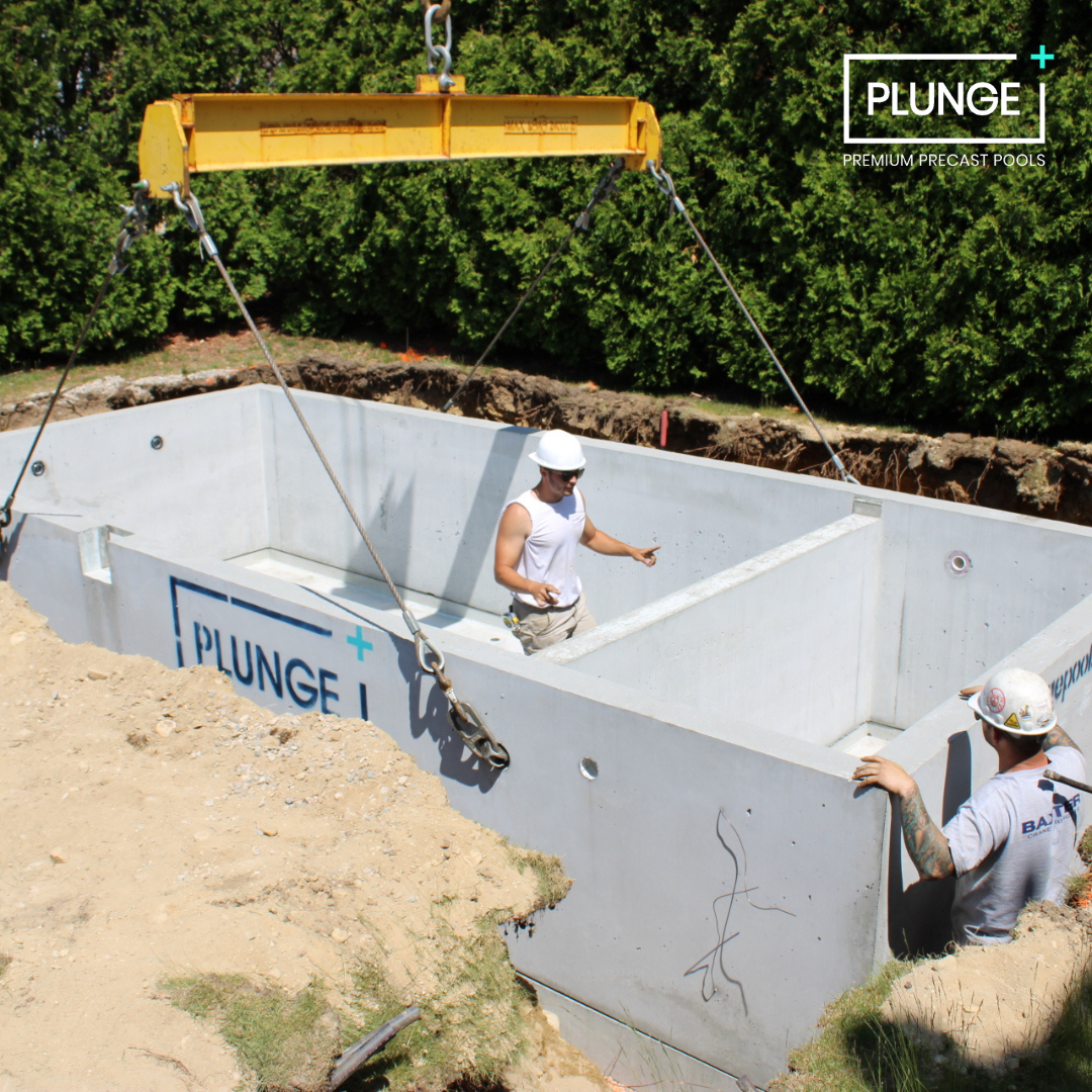 Construction workers install a Bulletproof pre-cast concrete pool called PLUNGE I at a construction site with dirt and green trees in the background.