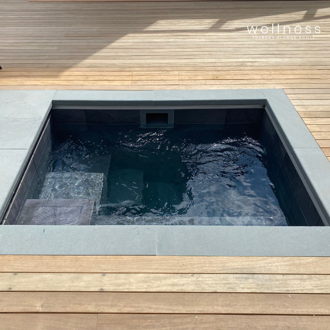 A small outdoor hot tub or spa with dark water, surrounded by light gray stone tiles and a wooden deck area.