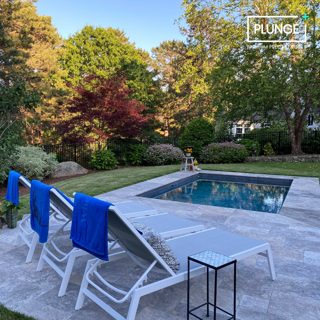 Backyard with a small swimming pool, four lounge chairs with blue towels, a small side table, and lush green trees and bushes in the background.