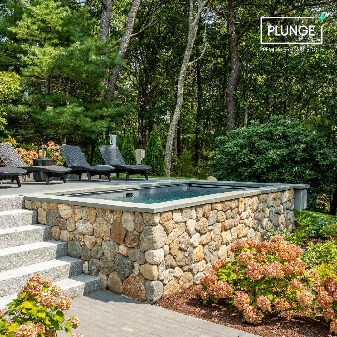 A backyard with a hot tub on a stone platform, surrounded by lush greenery and colorful flowers, with four black lounge chairs and trees in the background.