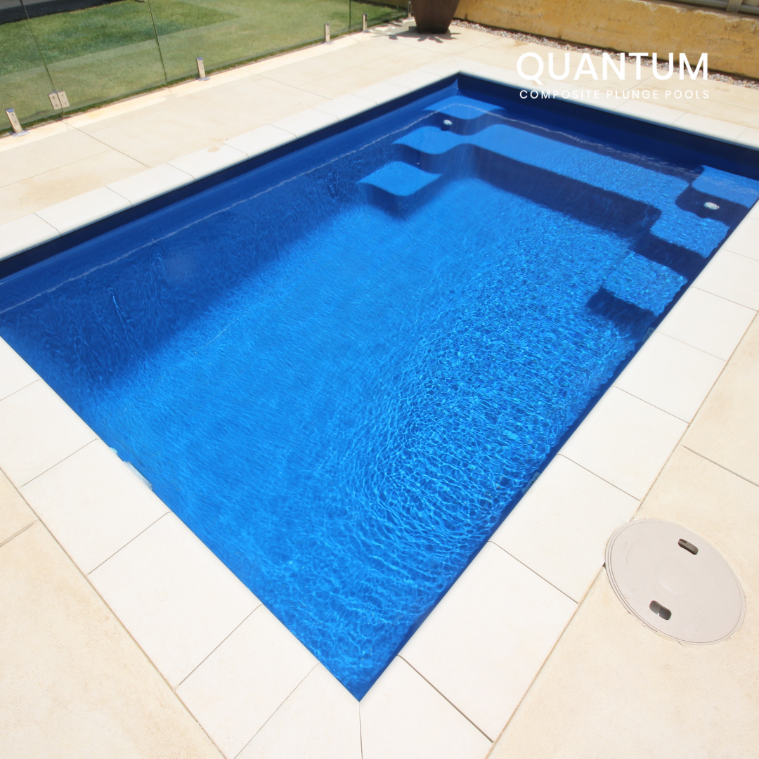 A modern blue rectangular pool with integrated steps, surrounded by a white tiled deck and glass fencing, with a potted plant in the background.