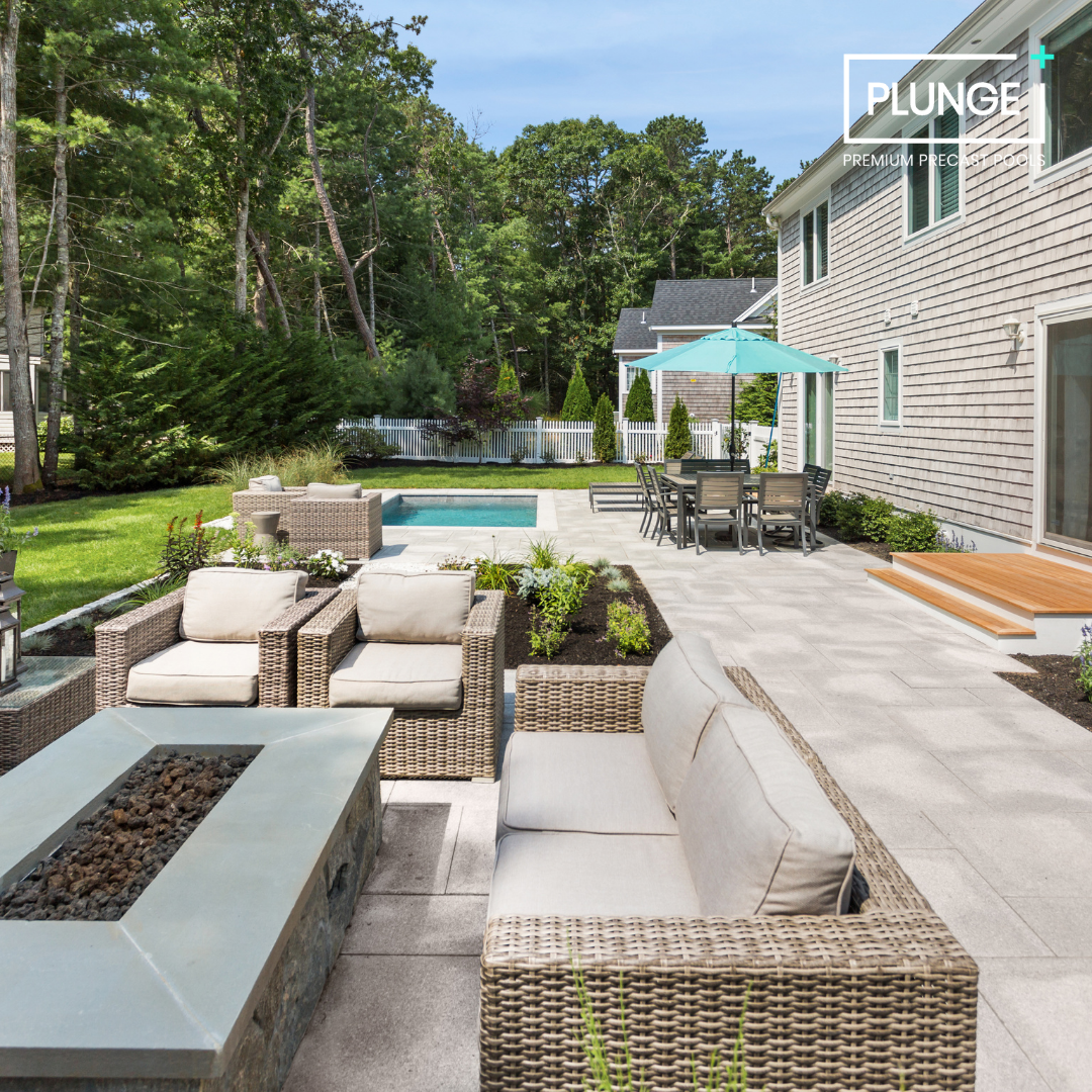 Backyard patio with wicker furniture, a fire pit, a swimming pool, a dining area with a blue umbrella, and garden with trees and bushes behind a white picket fence.