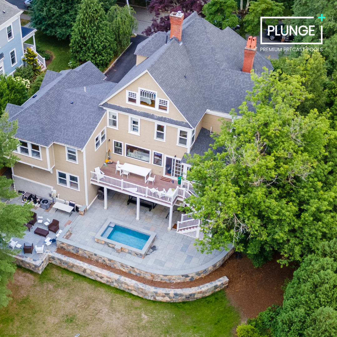 A large beige house with gray roofs, surrounded by green trees. There is a backyard featuring a hot tub, patio furniture, and a deck with outdoor dining. The house has multiple windows, a chimney, and a stairway leading to the yard. The photo is take