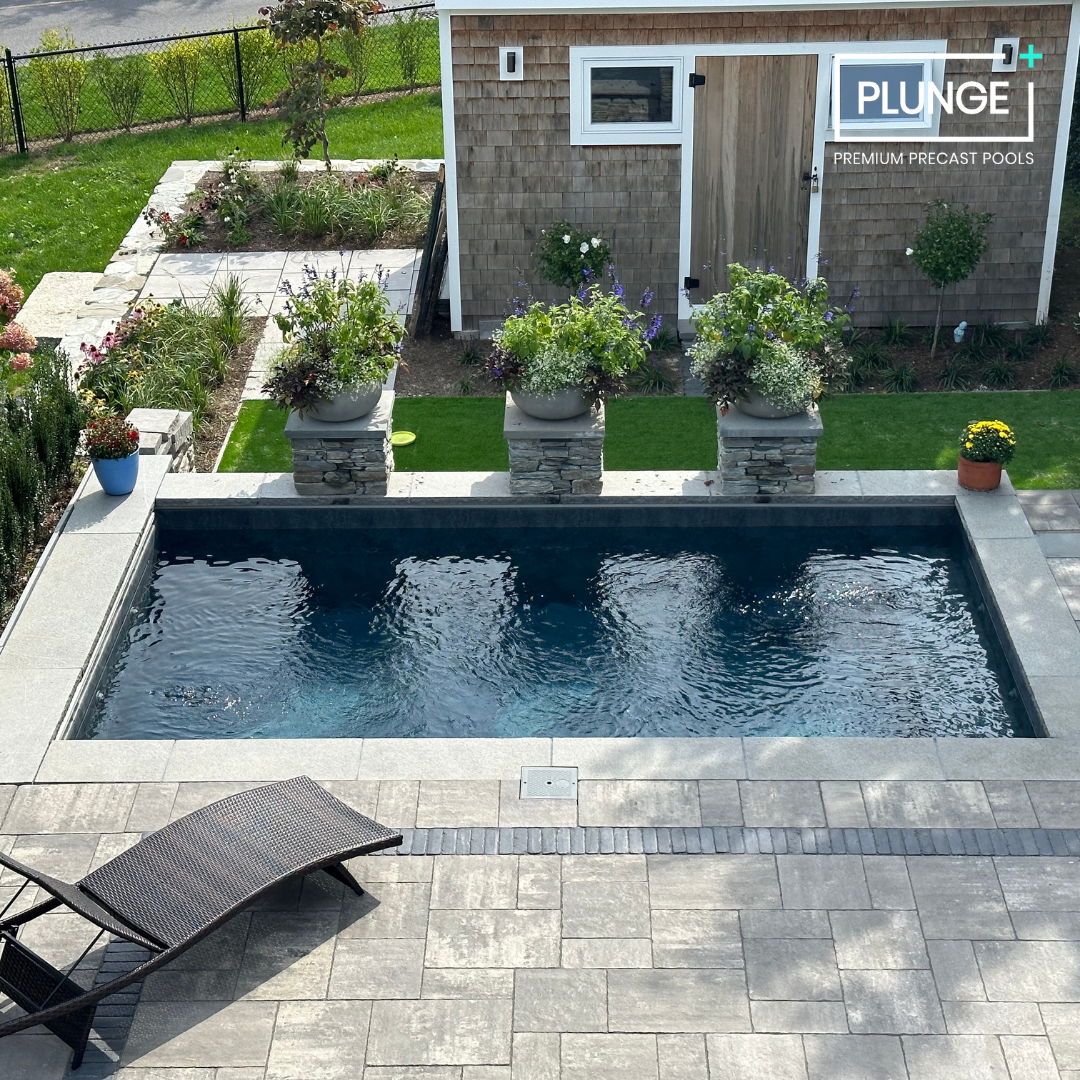 Backyard with a small in-ground swimming pool, lounge chair, potted plants, a shed with windows and a door, landscaped garden area with flowers and shrubs, and a grassy yard in the background.