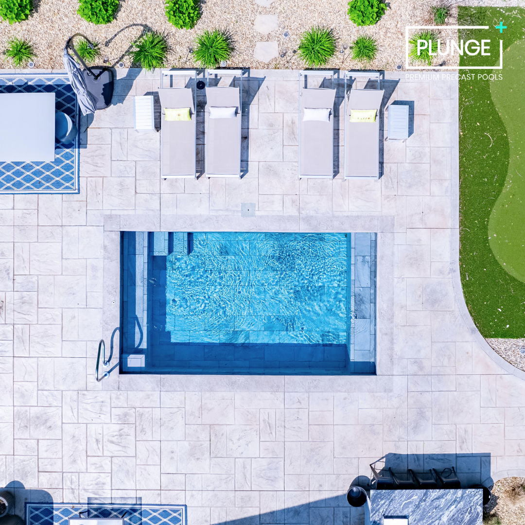 Aerial view of a swimming pool area with four lounge chairs, a small table, and a shaded seating area on a tiled patio, surrounded by greenery and a landscaped yard.