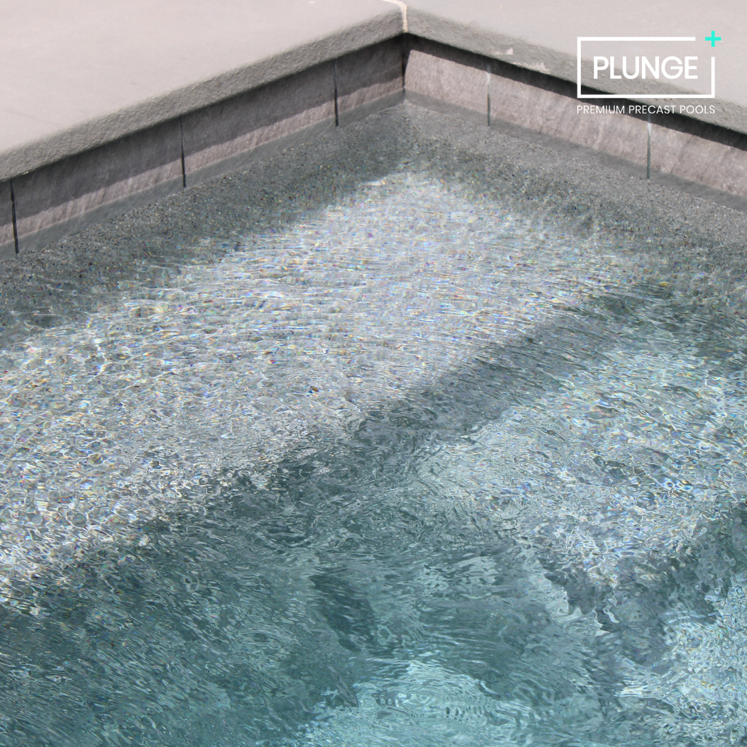 Close-up of a luxury swimming pool with clear water and textured pool edge tiles, with a logo in the corner that reads 'PLUNGE Premium Precast Pools.'