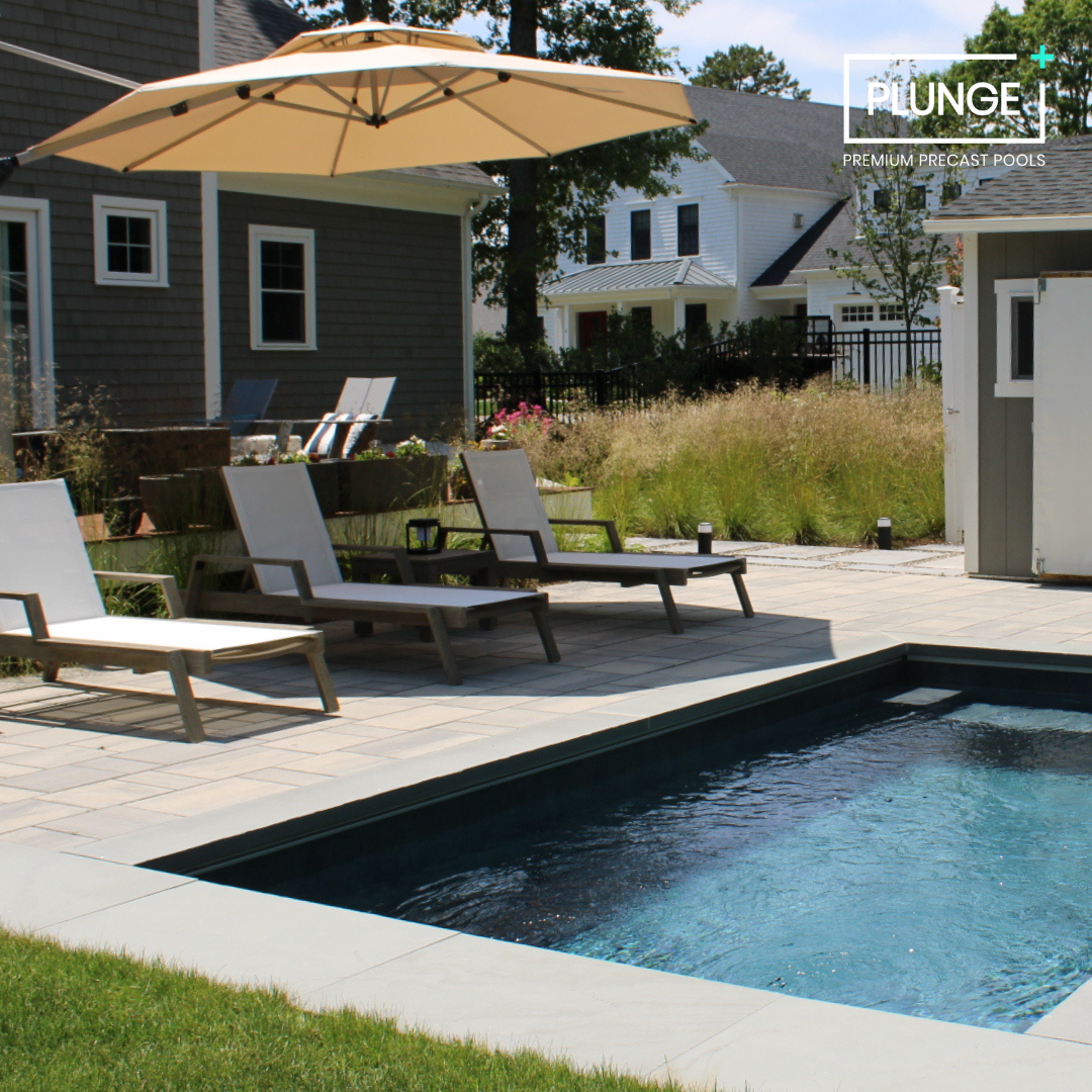 A backyard with a small in-ground swimming pool, lounge chairs, and a large beige patio umbrella. There are plants and grass surrounding the area and neighboring houses in the background. The image includes a logo for 'PLUNGE Premium Precast Pools'.