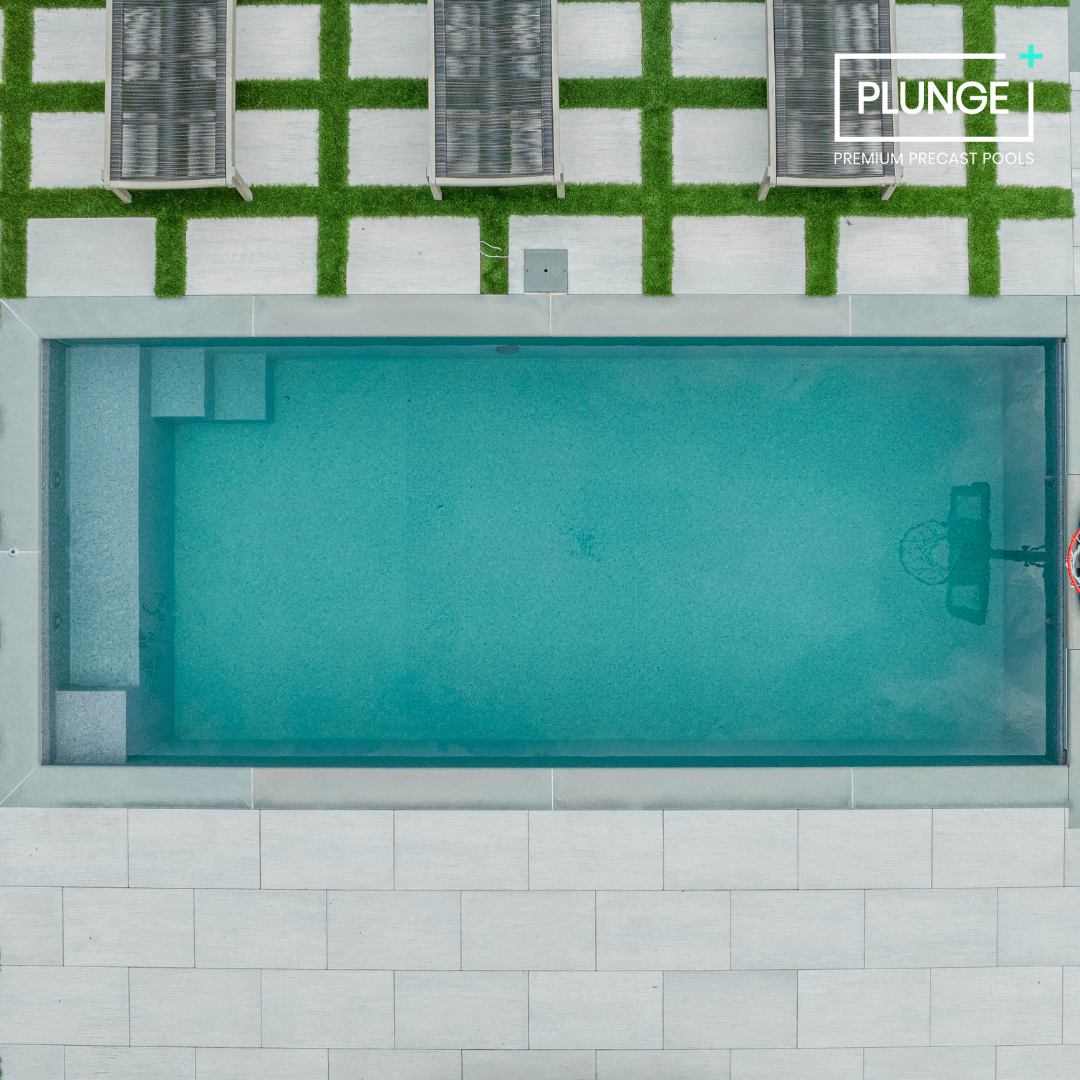 Aerial view of a rectangular in-ground swimming pool with a basketball hoop at one end, surrounded by white tiles and a patio with grass and deck chairs.