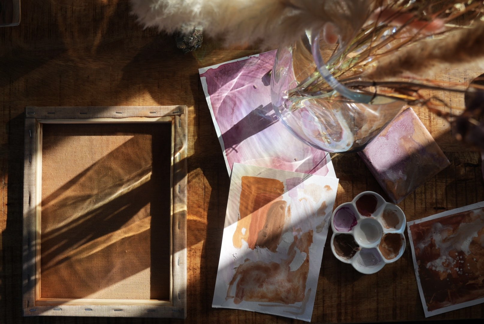 Ein Tisch mit Wasserfarben, Pinseln, Blättern mit Acryl- oder Aquarellfarben, ein leeres Holzbildrahmen, eine Vase mit getrockneten Blumen, stilvolles Arbeitssetup bei natürlichem Licht.