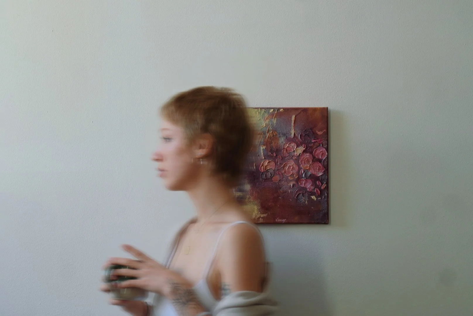 Eine junge Frau mit kurzen braunen Haaren hält einen Gegenstand in den Händen, während sie an einer weißen Wand mit einem bunten Blumenbild hängt.