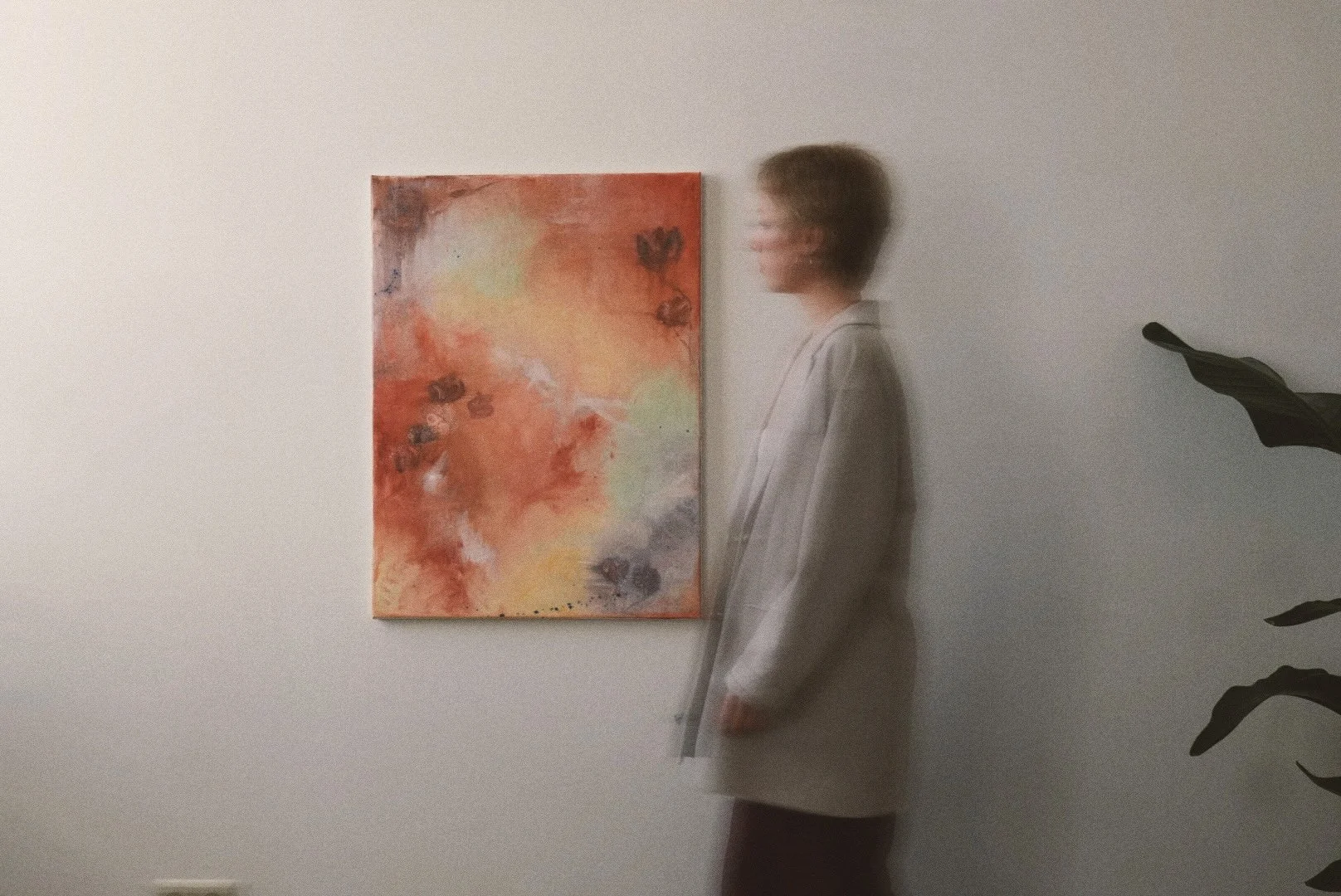 Eine Person in einem grauen Mantel steht vor einem abstrakten Gemälde mit warmen Farbtönen an einer weißen Wand.