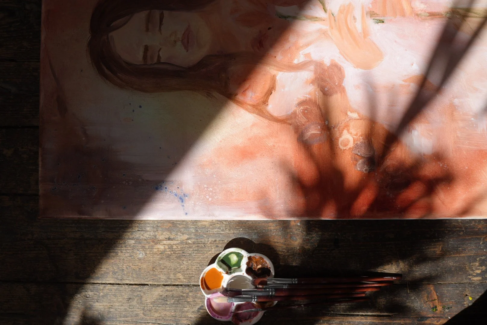 Unter einem Lichtstrahl befindet sich eine halb fertige Aquarellzeichnung eines nackt schauenden Frauenkopfes. Auf einem Holzuntergrund steht eine Farbpalette mit verschiedenen Farben und drei Pinsel.