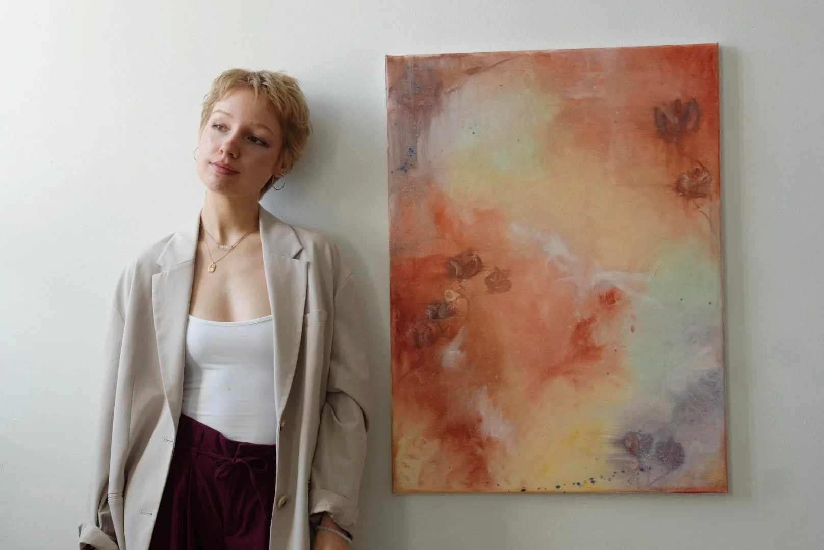 Eine junge Frau mit kurzen, blonden Haaren steht neben einem abstrakten Gemälde in warmen Orange- und Gelbtönen, das an einer weißen Wand hängt.