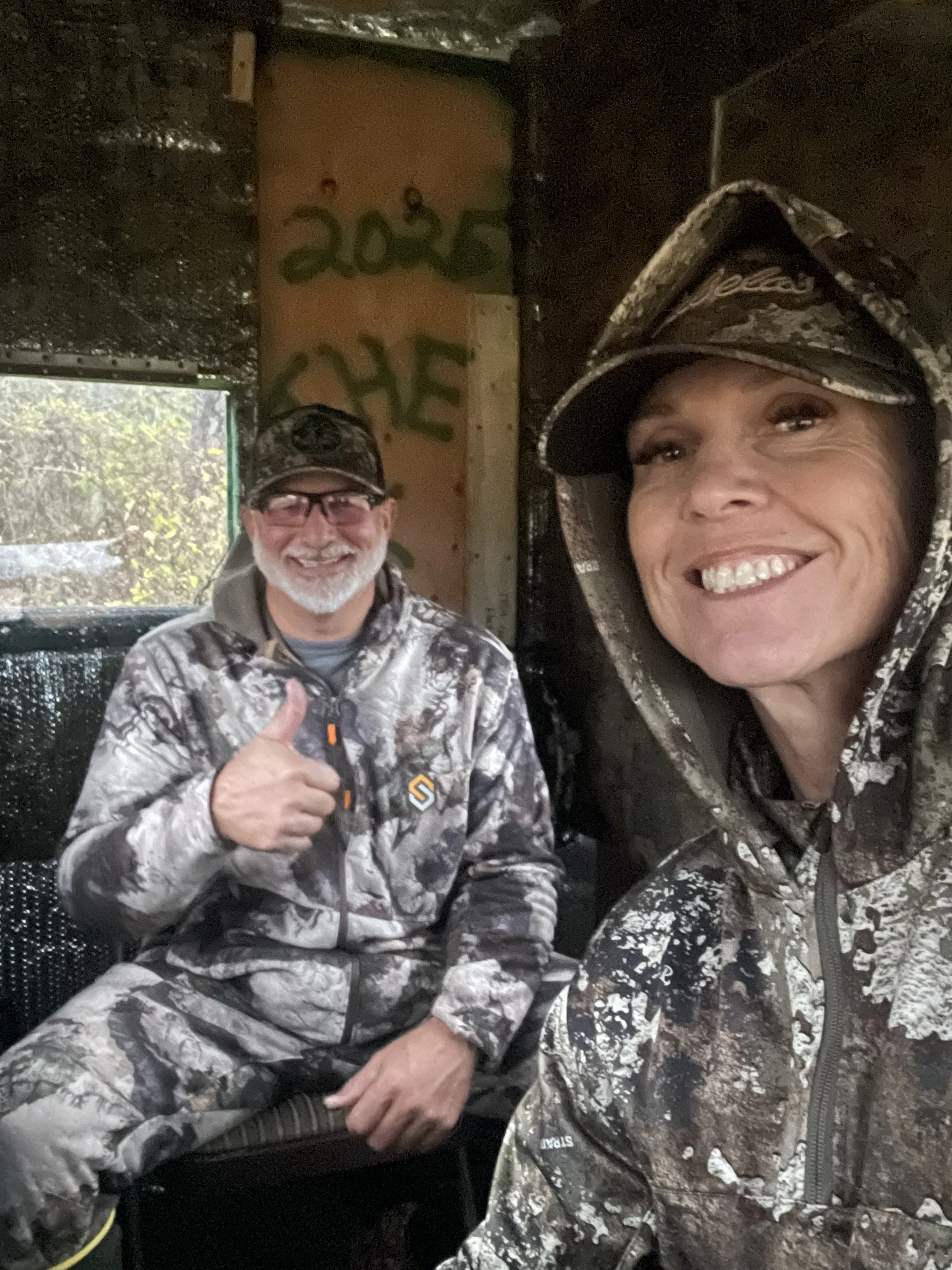 Two women in camouflage outdoor clothing taking a selfie inside a wooden, tree stand or hunting blind, with one woman giving a thumbs-up and both smiling.