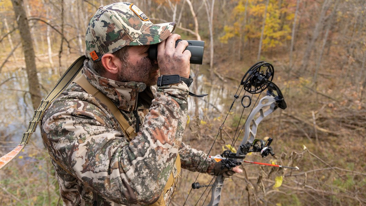 A man in camouflage clothing and a hat uses binoculars on a wooded riverbank, holding a compound bow in his other hand.