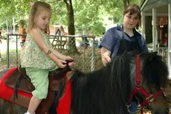 Two young girls, one riding a small black pony with a red saddle, and the other standing beside it, outdoors with trees and a building in the background.