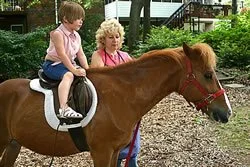 A young girl riding a small brown horse with a woman walking beside them in an outdoor area.