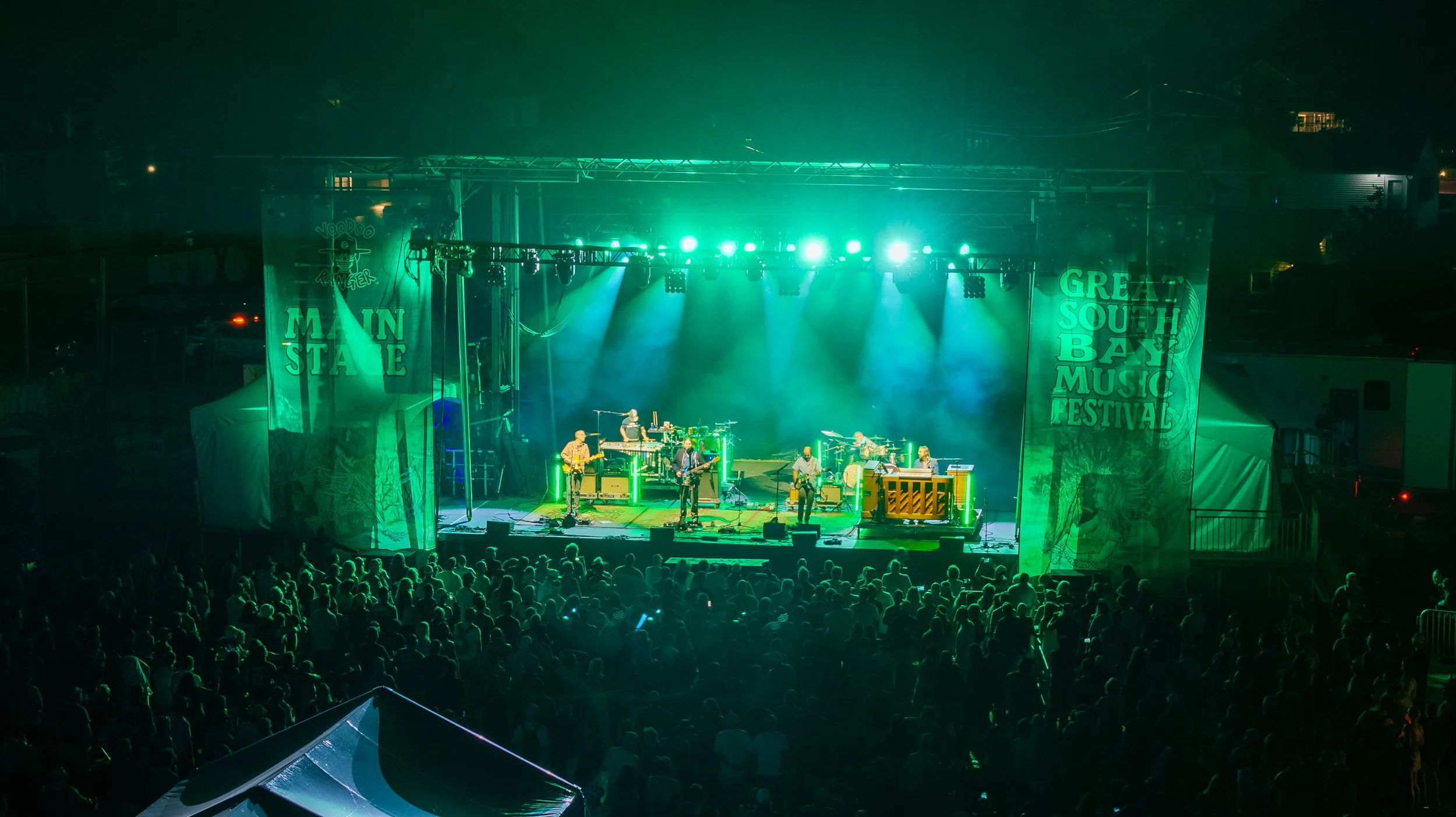 Live outdoor music concert at night with stage illuminated in green, band performing, and an audience gathered in front.