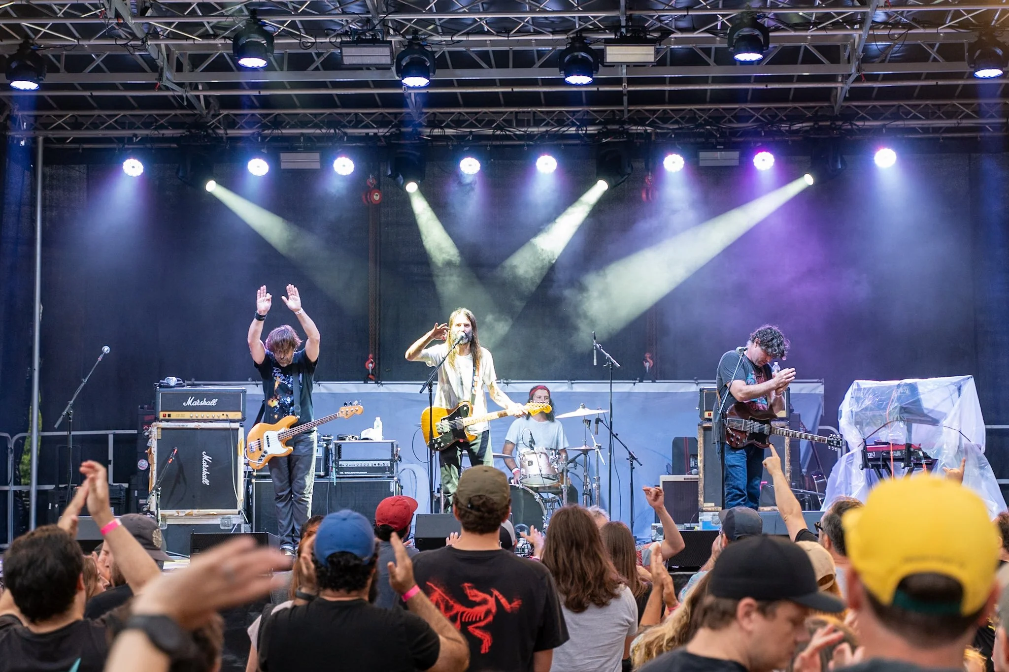 A band performing on an outdoor stage with four members, including a drummer, a guitarist, and a bass player, under colorful stage lights, surrounded by an audience.