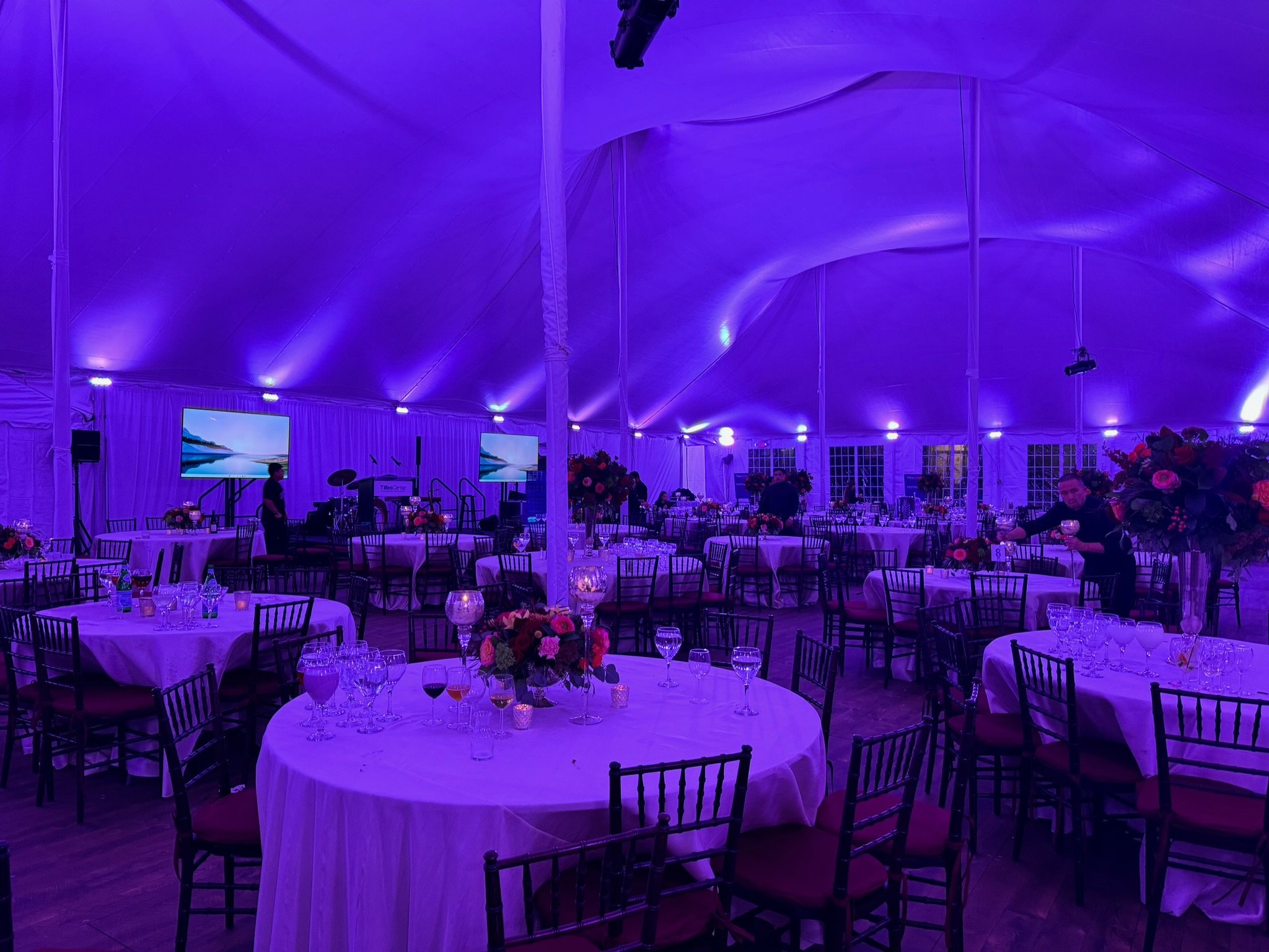 Event space decorated with purple lighting, round tables with white tablecloths, flower centerpieces, and glassware, with a stage, musical instruments, and two large screens in the background.