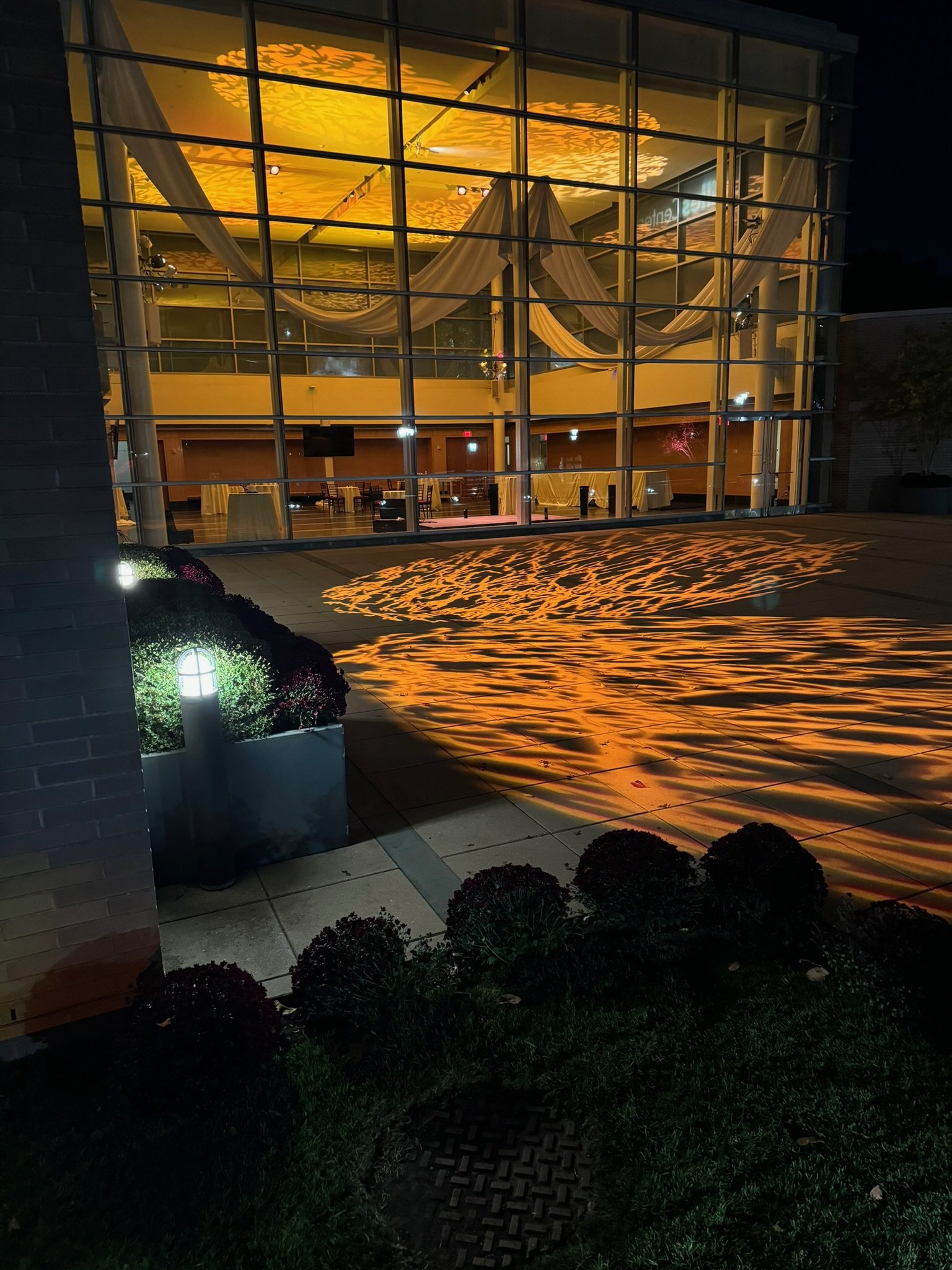 Nighttime view of a building with large glass windows, decorated with drapes and warm yellow lighting. The reflection of the lighting creates a pattern on the ground outside.