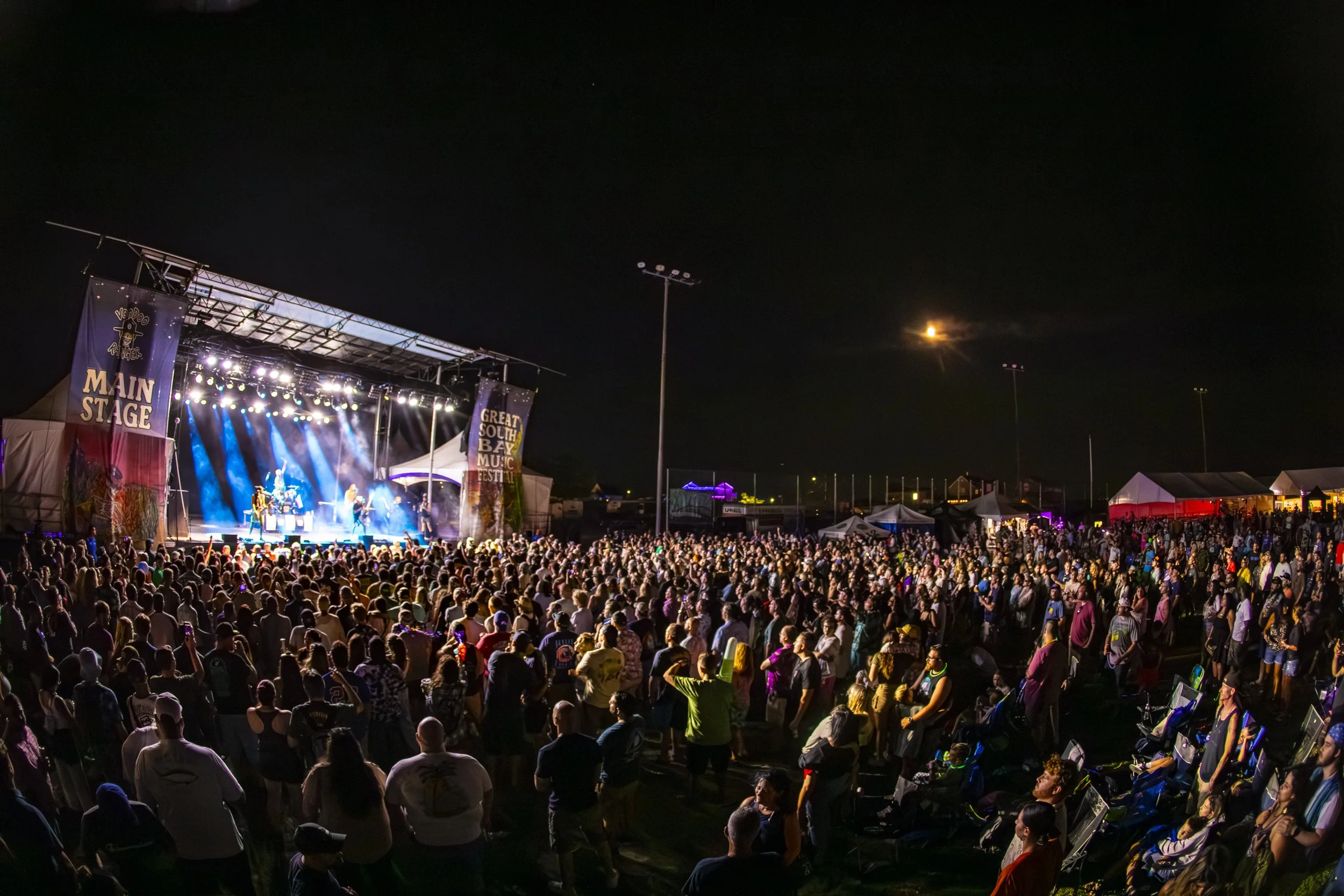 Crowd of people watching a live concert on an outdoor stage at night, with bright lights and tents in the background at the Great South Bay Music Festival.